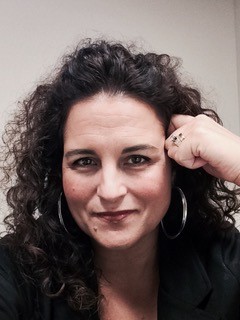 Kristen is a white woman with dark brown curly hair. She is posed with a curled hand resting against her right temple, and is wearing a black blouse.