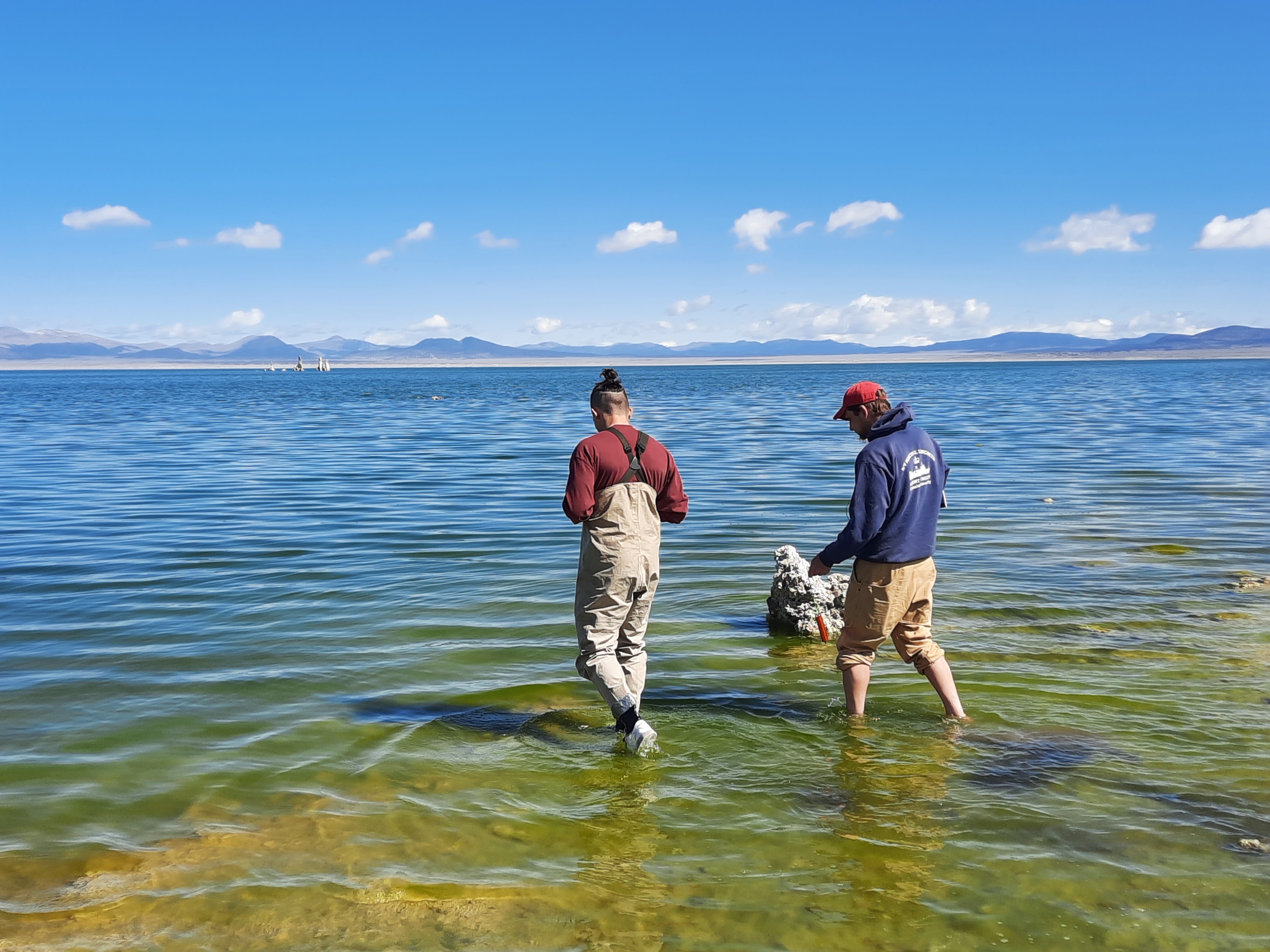Two people wade ankle deep into a body of water. Wide blue sky over the horizon. 