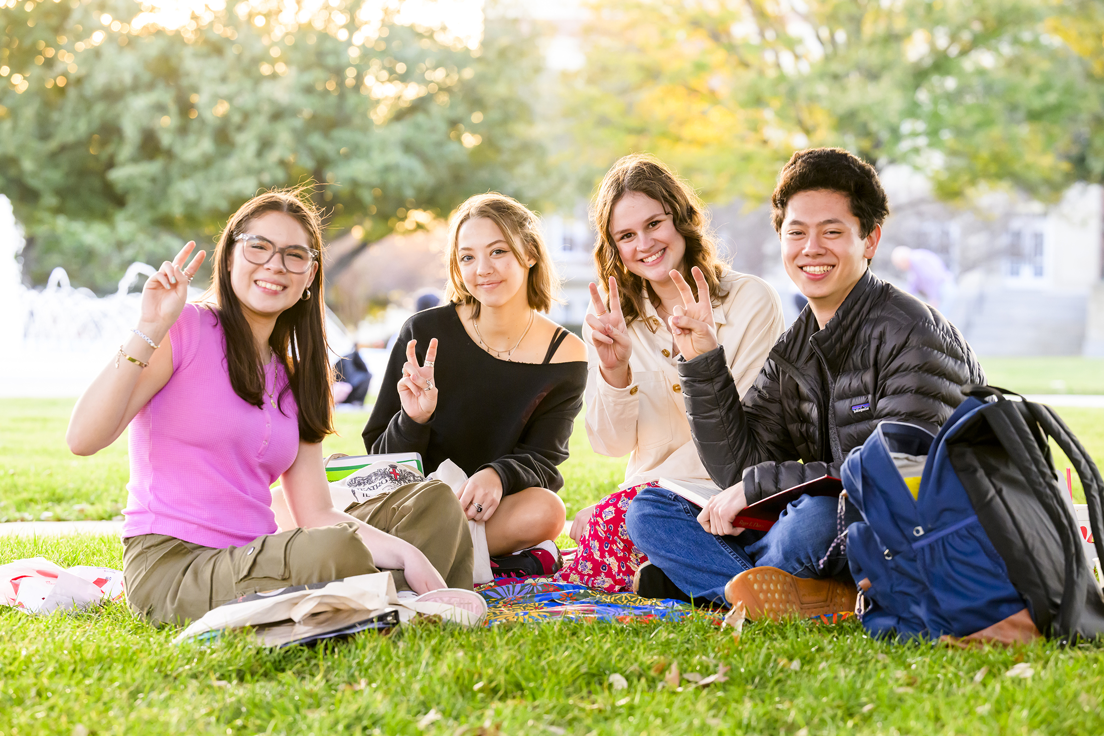 Four students are relaxing on the grass at SMU, smiling and holding up their fingers in the University's traditional "pony up" greeting.