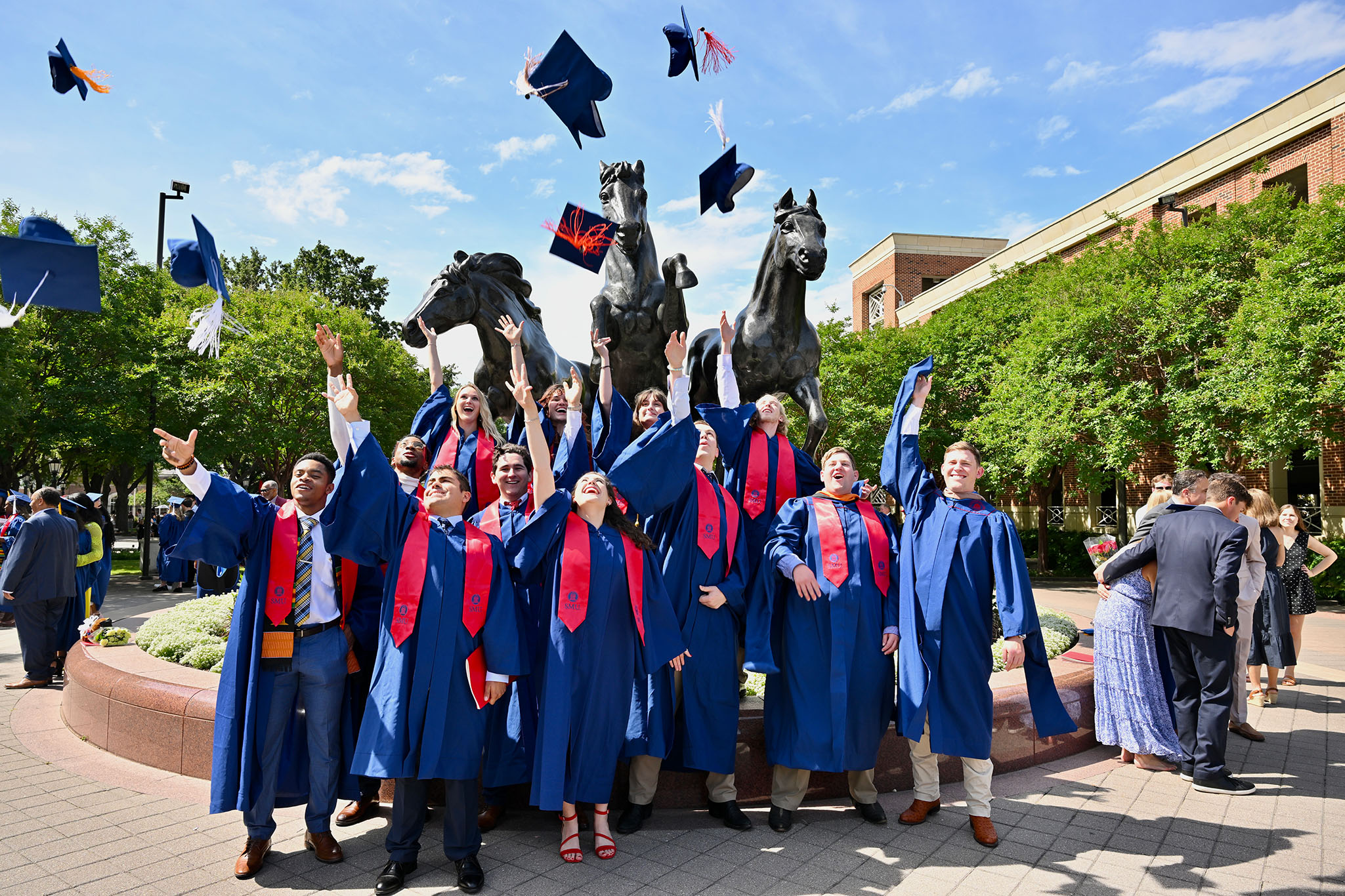 students in graduation regalia tossing caps in the air in front of mustang statue