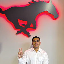 Zidan Thomas makes the pony ears hand sign in front of a wall with the SMU logo