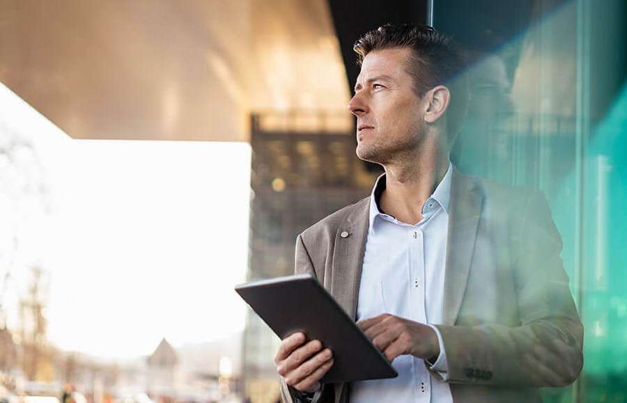 A male business administrator outside of an office building, holding a tablet, and looking into the distance.