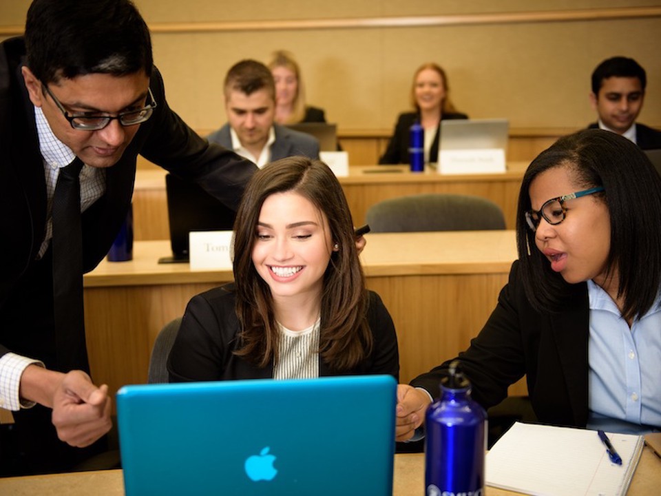 A lecture hall full of students working on laptops with a faculty member assisting 