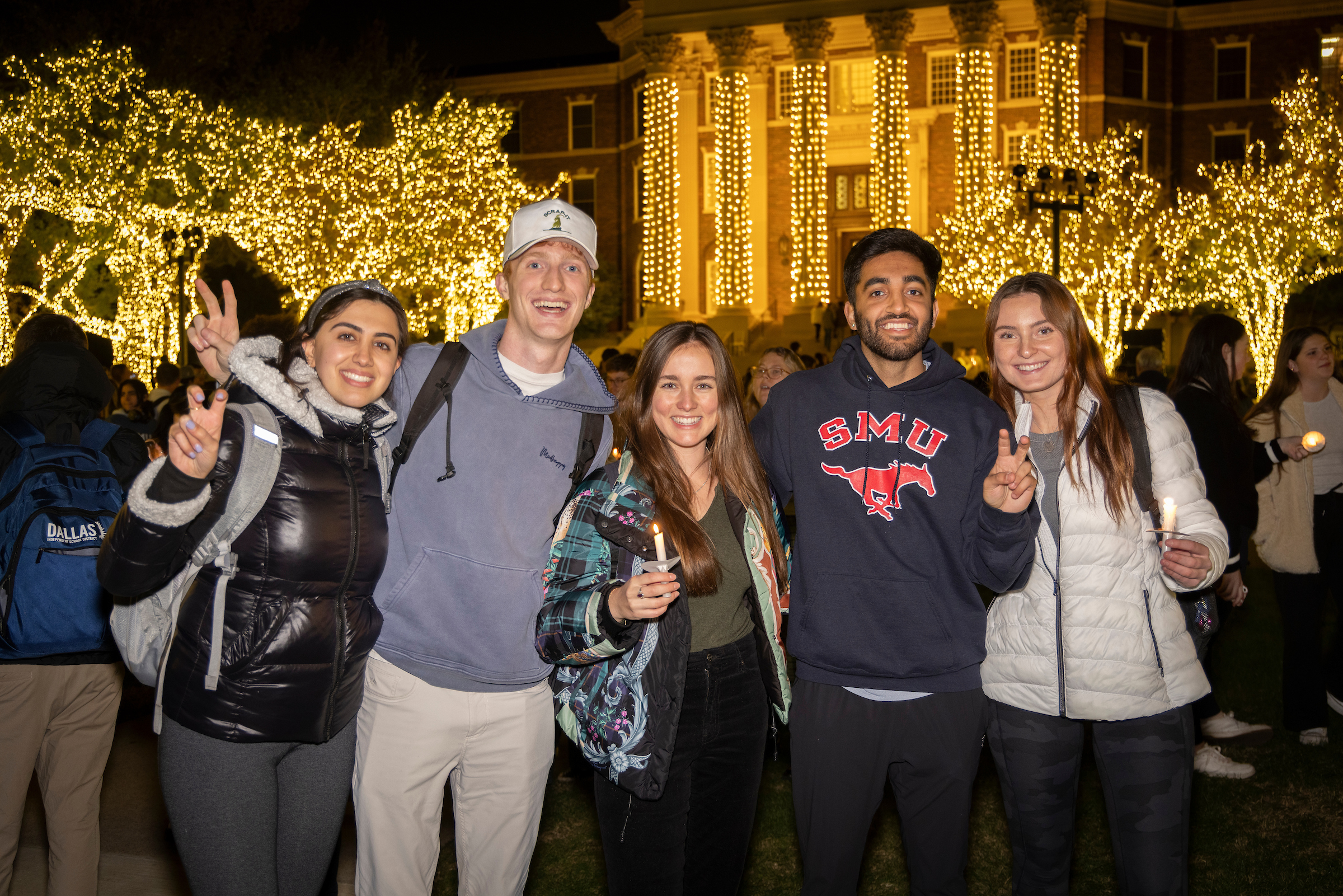 Three female and two male students, dressed warmly and smiling, stand in front of Christmas lights on the SMU campus. They are all smiling and several are making the pony ears hand gesture with their index and middle fingers in a V shape, slightly bent