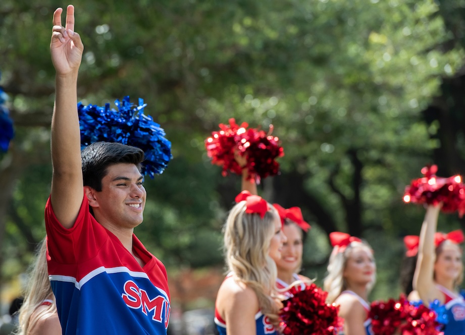 A male cheerleader, wearing a red and blue short-sleeved SMU uniform, smiles as he raises his right arm and makes the "pony ears" hand gesture with his index and middle finger raised and slightly bent, on a sunny day on campus. Several female cheerleaders are nearby.