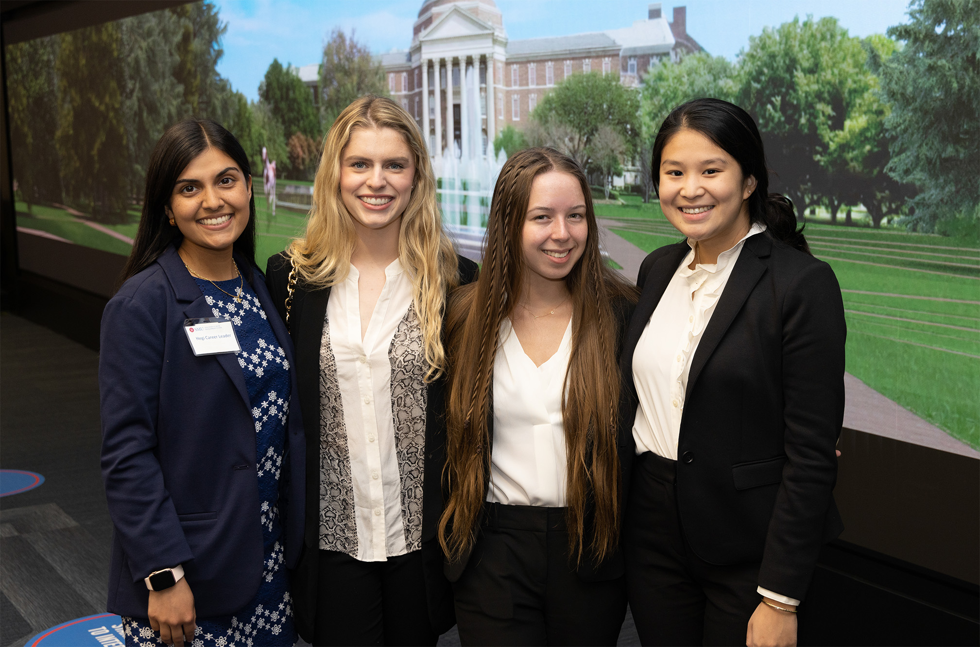 four students standing in front of video wall smiling at camera