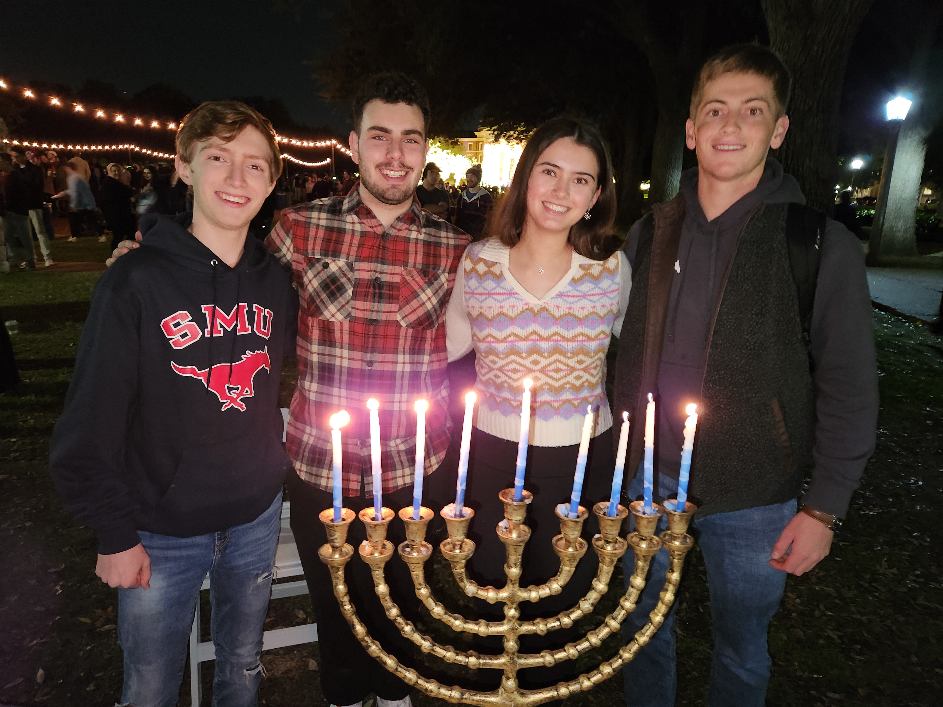 students standing behind a menorah with seven white candles all lit outdoors at night