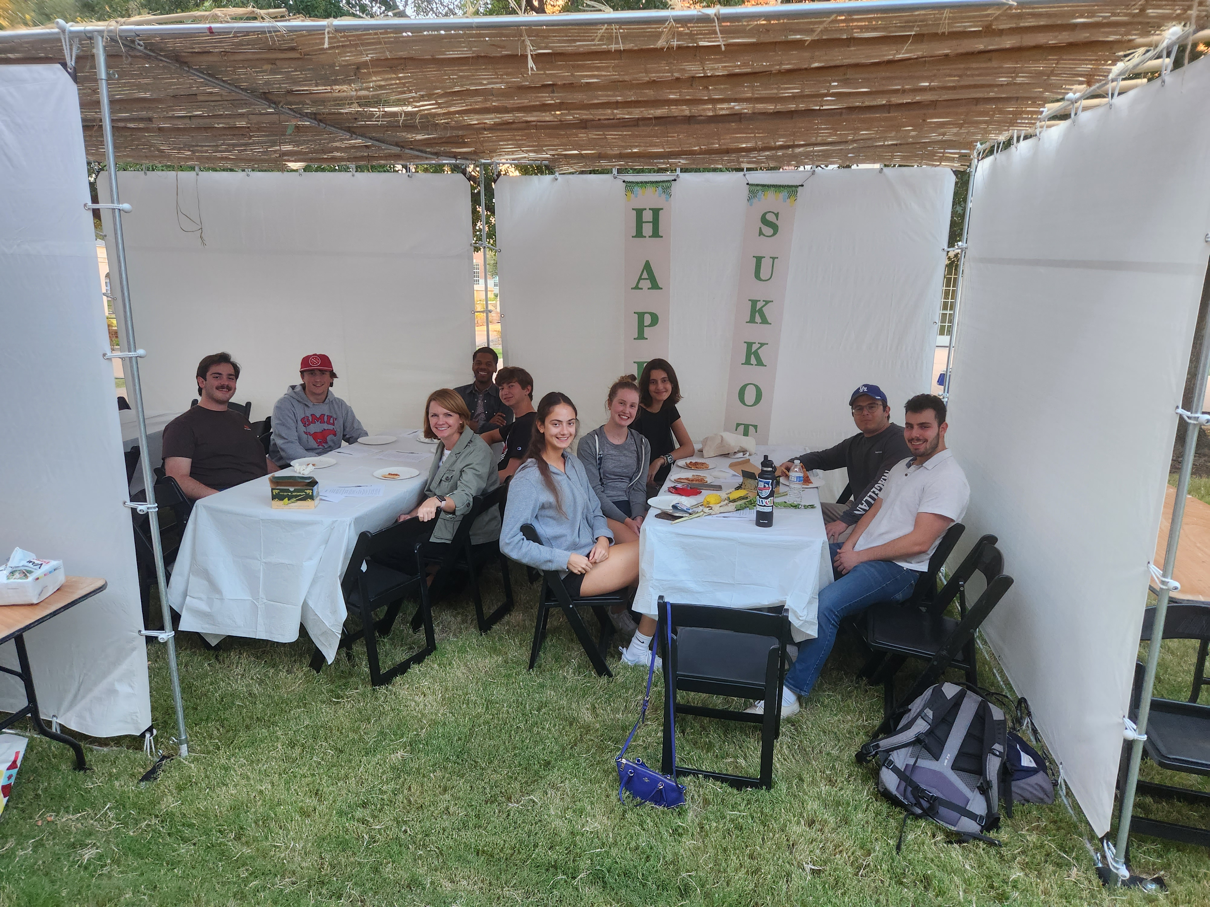 students at tables outdoors on grass surrounded by white drapes covered with string lights