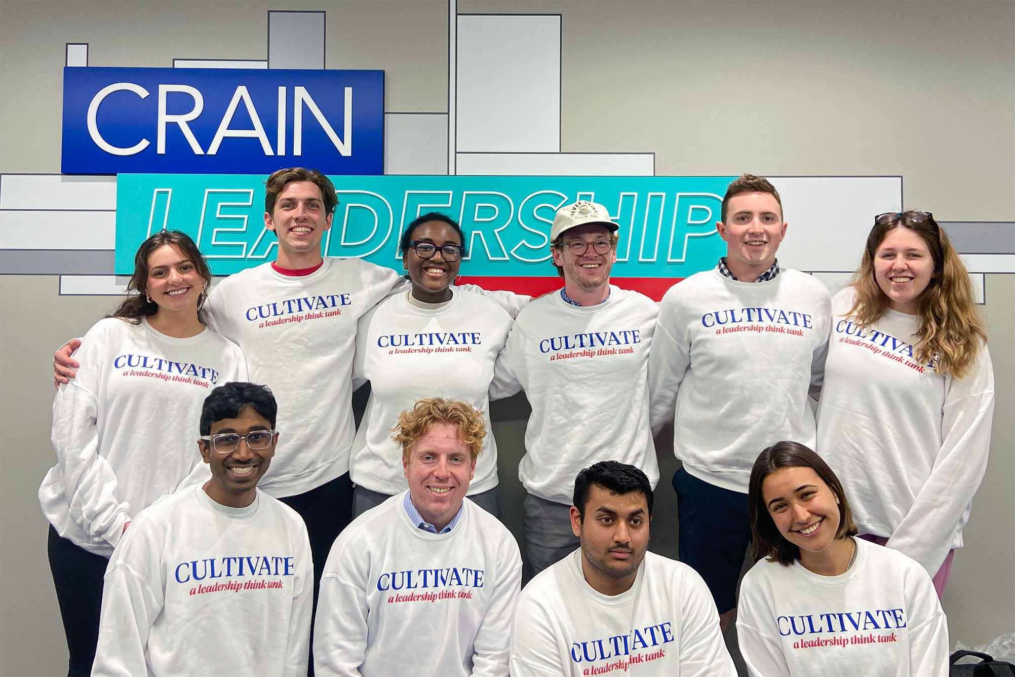 group of students in white sweatshirt with "Cultivate" in blue ink and "a leadership think tank" in red ink, standing in front gray wall of sign "Crain Leadership"