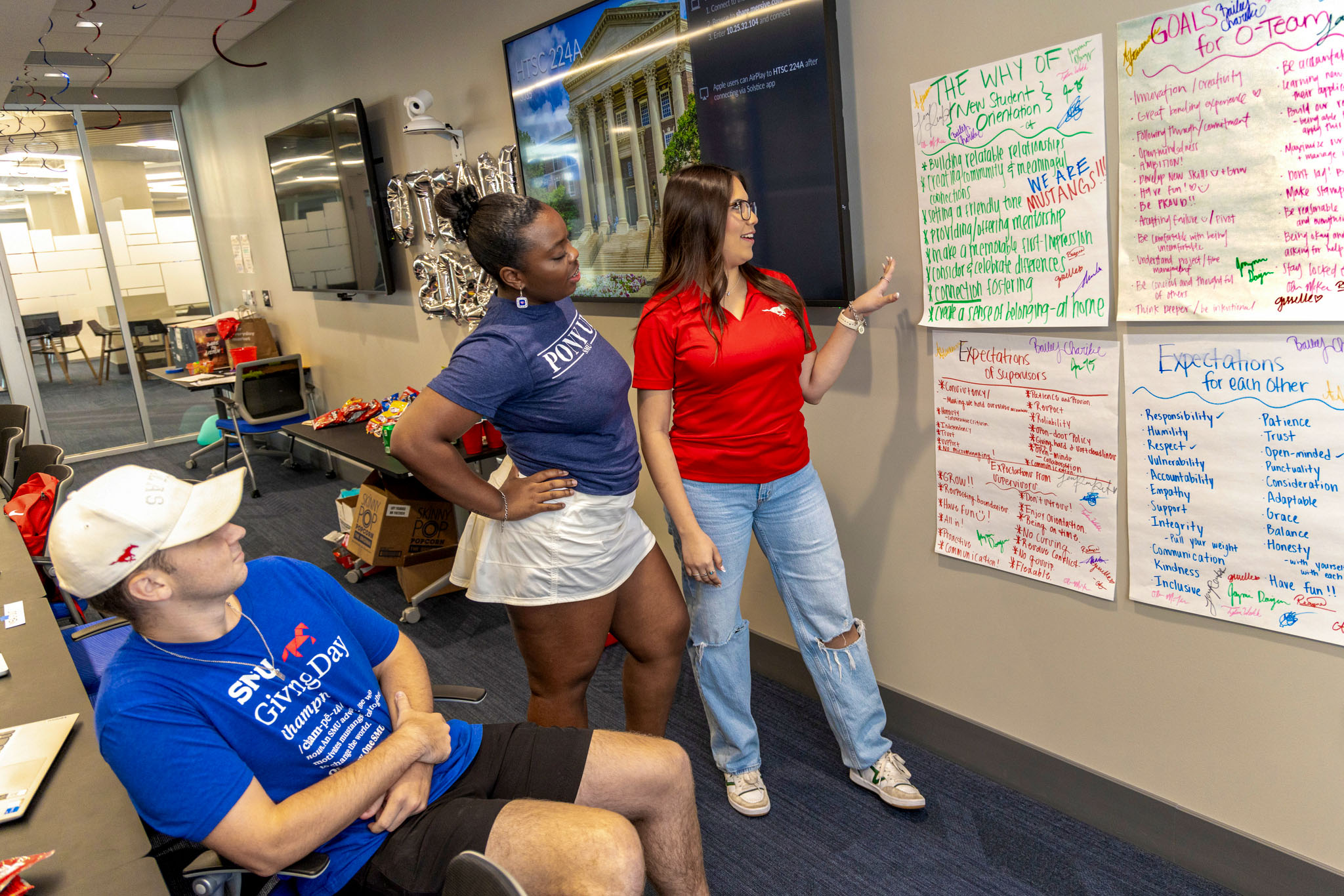 three students wearing red and blue shirts talking and looking at work hanging on the wall