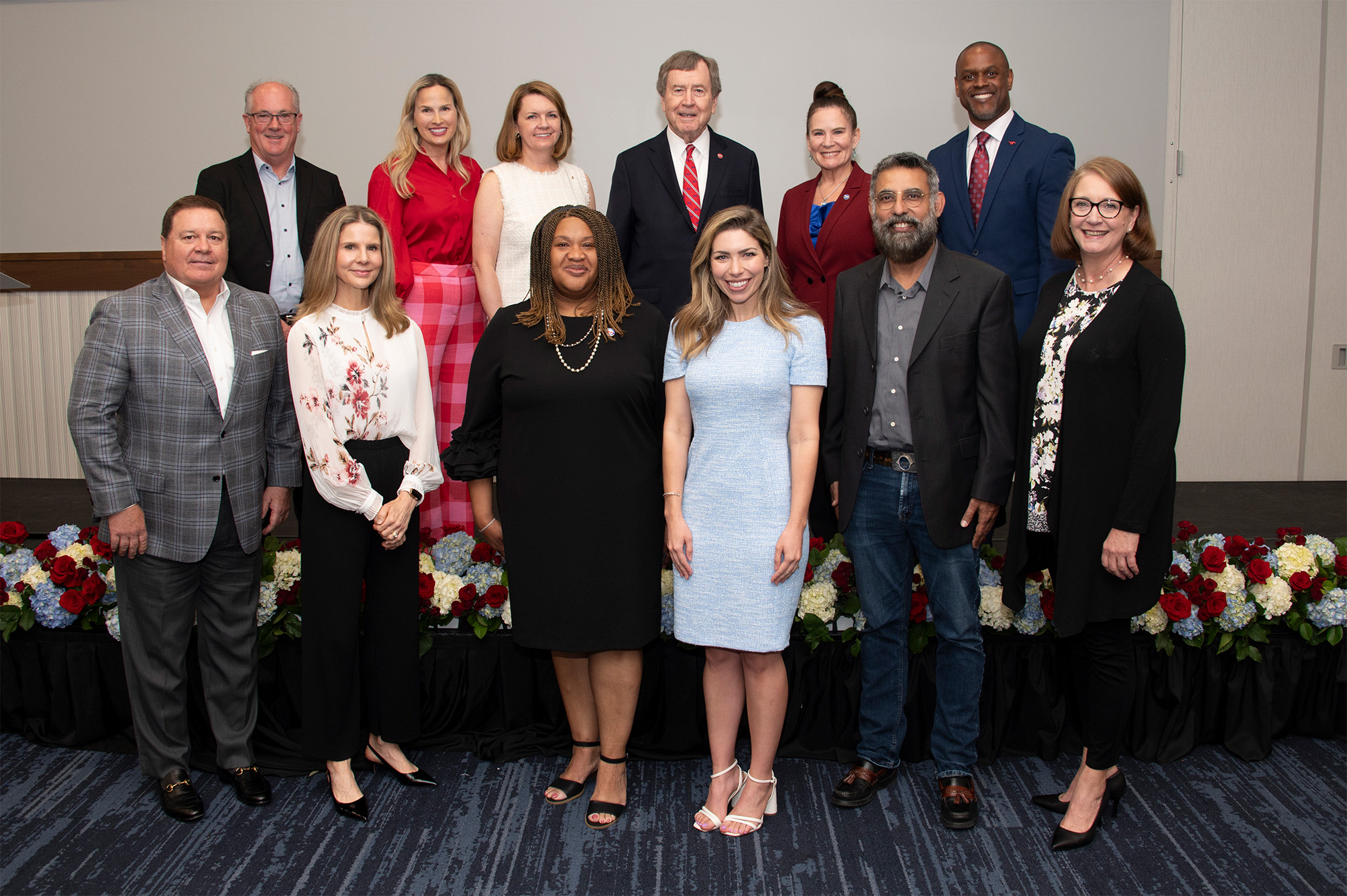 group of people standing in two rows, back row is standing on platform, in front of gray wall with red, white, and blue flowers on ground