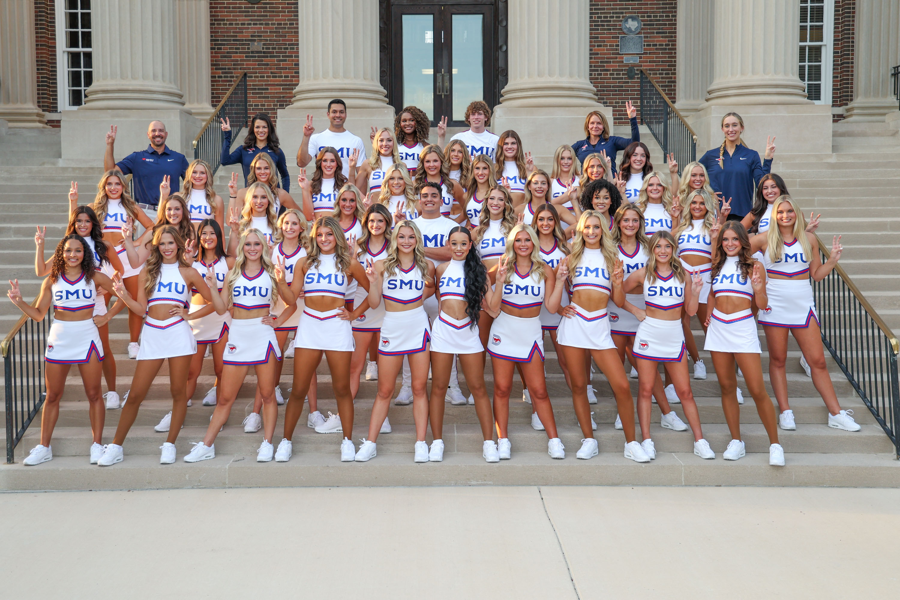 2024-2025 full cheer and pom team photo, standing outside on stairs in front of building in white uniforms, coaches standing with team in black SMU polo shirts