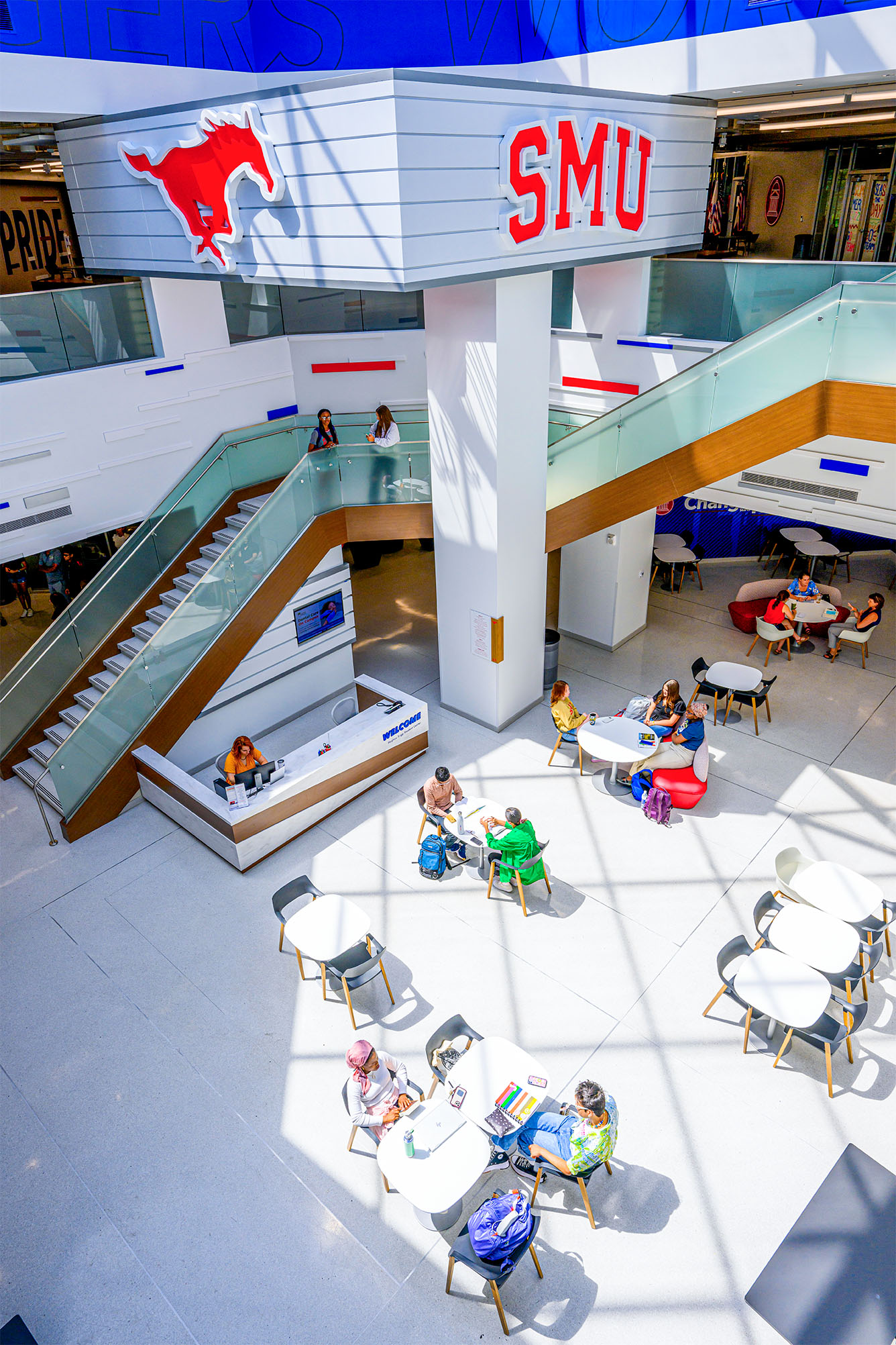 indoor atrium of building with students sitting at tables, a large staircase to the next floor, a red mustang cutout and SMU in red hanging on wall